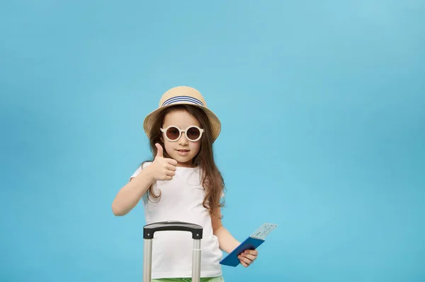 Isolated portrait on blue color background of a little traveler child girl in sunglasses and straw hat, holding a flight ticket, thumbing up, going for weekend getaway, isolated blue color background