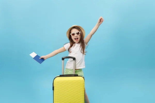 Cute little girl in summer wear, straw hat and sunglasses expressing happiness while looking at camera while holding passport and boarding pass while standing with yellow suitcase over blue background
