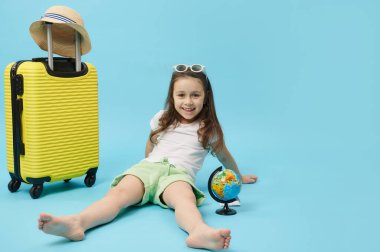 Smiling Caucasian adorable child traveler tourist adventurer, cute mischievous little girl rejoicing while going for summer getaway, isolated blue color background, near a yellow suitcase and globe