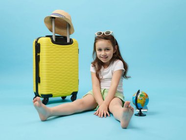 Charming Caucasian 5 years old little girl sitting barefoot on blue background, near a yellow suitcase and globe, cutely smiling looking at camera. Traveler tourist child going for summer holidays