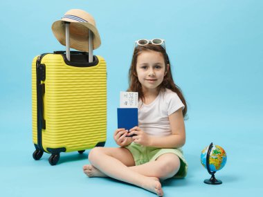 Adorable traveler kid, cute little girl in summer wear, smiles looking at camera, holds a flight ticket and passport, sitting near globe and yellow suitcase on blue background. Trip and travel concept
