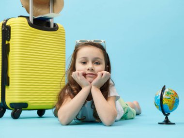 Close-up portrait Caucasian adorable little traveler child girl, with globe and yellow suitcase, looking at camera isolated on blue color background. Tourism around the world. Trip and travel concept