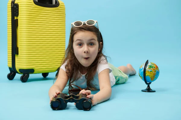 Happy and surprised little traveler child girl with binoculars, expressing positive emotions and surprise, looking at camera, lying near a yellow suitcase and globe, isolated on blue color background