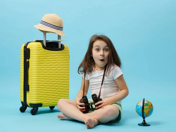 Surprised little girl with binoculars in her hands, sitting barefoot near yellow suitcase and globe, expressing surprise looking at camera, isolated on blue background. Adventure and travel concept
