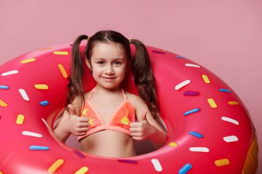 5 yaşında sevimli bir beyaz çocuk, parlak mayo giymiş şirin küçük bir kız, başparmakları havada, kameraya gülümseyen, pembe arkaplan üzerinde şişirilebilir bir donut yüzme halkasının içinde poz veren
