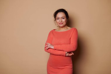 Confident portrait of a beautiful multi-ethnic middle-aged woman, successful entrepreneur, sales manager in orange casual dress, looking at camera, posing with folded hands over beige background
