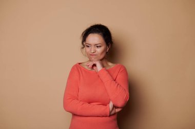Frustrated middle-aged woman in orange dress, expressing confusion while reasoning, isolated over beige color background