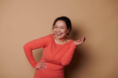 Happy dark-haired multi-ethnic woman in stylish orange dress, smiling a beautiful toothy smile looking at camera, expressing positive emotions, laughing isolated on beige color background
