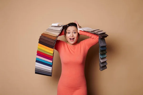 Beautiful woman with fabric swatches expressing surprise by a large assortment of fabric of various colors and textures, isolate on beige background. Room renovation. Home improvement.Interior design