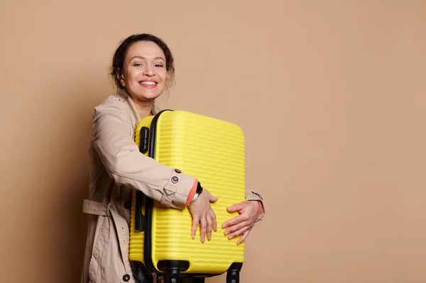 Beautiful dark-haired woman passenger, wearing a stylish beige coat, hugging her yellow suitcase, smiles looking at camera, isolated over beige background. People, journey, trip, travel abroad concept