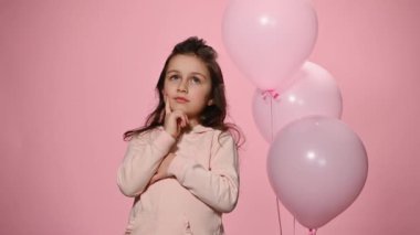 Beautiful little child girl, holding finger on her chin while thoughtfully looking aside, smiling looking at camera, expressing idea, isolated on color background with inflatable pink pastel balloons