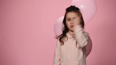 Mutlu çocuk, pembe pastel renkli balonları olan doğum günü kızı, bir iki üç tane sayıp pembe arka plandaki reklamlar için fotokopi alanını gösteriyor, kameraya gülümsüyor, başparmağıyla işaret ediyor.