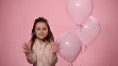 Lovely cheerful little girl in pink pastel wear, clapping her hands and jumping against a color background with inflatable helium balloons, wishing you a happy birthday and smiling looking at camera