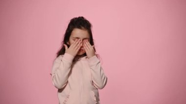 Charming lovely little child covers her eyes with hands till the helium balloons flying over pink pastel color background. Cheerful baby girl rejoices looking at camera while balloons cannot take off