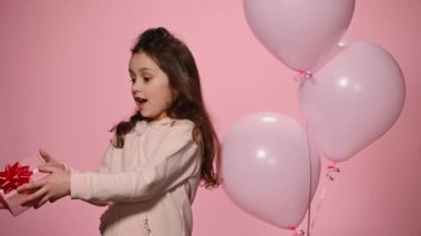 Adorable little child girl in pink sweatshirt, receives a cute gift box with happy surprise for her birthday and shows it to camera, standing near pink helium balloons over isolated color background.