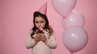 Pembe şenlik şapkalı, doğum günü pastası ve şişme helyum balonlarıyla pembe arka planda poz veren beyaz bir kız. Doğum gününü kutlayan 5 yaşında bir çocuk.
