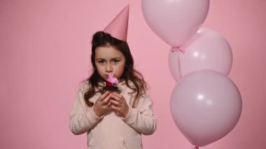 Caucasian lovely little girl in pink festive cap, posing on pink color background with a birthday cake and inflatable helium balloons. Happy 5 years old child celebrating her birthday