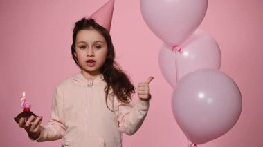 Happy little birthday girl celebrating her birthday party, making cherished wish, blowing out candle and expressing happiness and joy on isolated pink background decorated with helium pink balloons