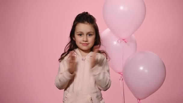 少女は カメラを見て笑みを浮かべて 孤立した色の背景にピンクのパステルインフレータブル風船の横にポーズ彼女の手を拍手 チャーミング陽気な子供があなたを迎え あなたに幸せな誕生日を願っています — ストック動画