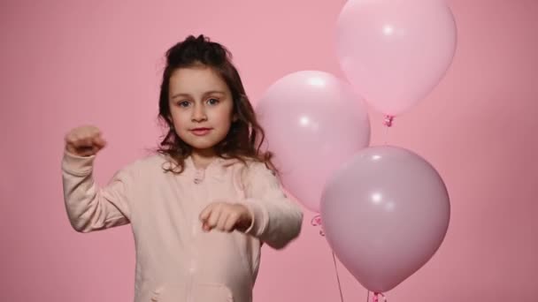 美しい長い巻き毛を持つ陽気な白人の少女は ピンクの服を着て 色の背景の上に音楽に移動し ピンクのパステルヘリウム空気風船の近くに立っている かわいい子供が楽しい — ストック動画