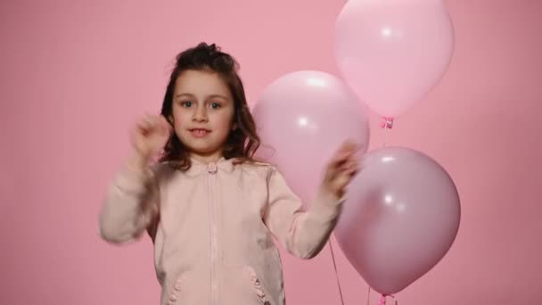 陽気な白人の小さな子供の女の子は ピンクのパステルの服を身に着けています 楽しみを持っています フリースタイルのダンスの動きを実行します インフレータブルヘリウム風船の近くに踊ります 隔離されたピンクの色の背景 — ストック動画