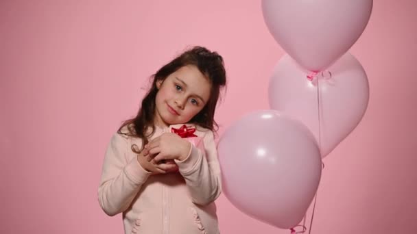 かわいい女の子のピンク色の背景に肖像画 優しく カメラで笑みを浮かべて 膨張ヘリウム風船の近くに立って 幸せな驚きとかわいいギフトボックスを抱いて 誕生日おめでとう — ストック動画
