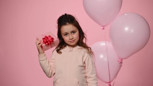白人5歳の素敵な女の子 ピンクの色の背景の上に立って ギフトボックスを振る 彼女の誕生日のためにそれの中の幸せな驚きを推測しようと ヘリウムピンクの風船の近くに立って — ストック動画