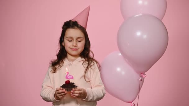 白人幸せな子供女の子でピンク誕生日キャップ 作る大切な願いと吹いてキャンドルオン彼女の誕生日ケーキ 近くに立って美しいインフレータブルヘリウム風船で隔離されたピンク色の背景 — ストック動画
