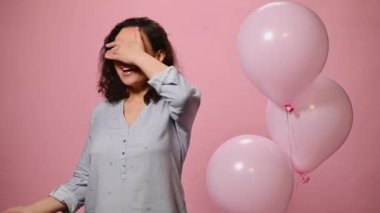 Charming woman covers her eyes, expressing surprise while receiving gift box with happy surprise for birthday or Womens Day, smiling at camera, on pink color background with inflatable helium balloons