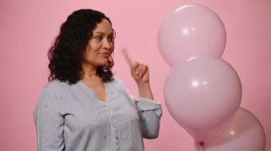 Mutlu çok ırklı orta yaşlı, mavi gömlekli güzel bir kadın, şişme helyum pembe balonlara dokunuyor, sevimli dişlek bir gülümsemeyle kameraya bakıyor, pembe arka planda poz veriyor.