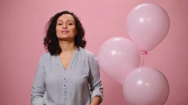 Beautiful attractive ethnic woman showing at camera a pink heart shaped gift box with a happy surprise for Saint Valentines Day, posing over a pink color background with inflatable helium balloons