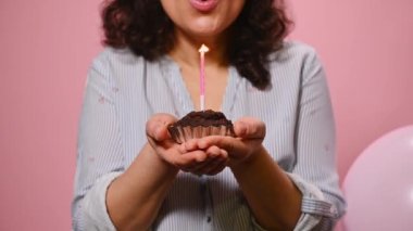 Selective focus on a delicious sweet chocolate cupcake in the hands of smiling birthday girl, blowing candle on pink color background with inflatable helium balloons. Woman celebrating birthday party