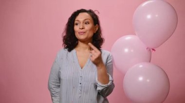 Attractive Hispanic woman shows silence sign, winks at camera and points with index finger at cute bouquet of chrysanthemum and gypsophila flowers, isolated on pink background with helium balloons