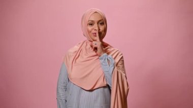 Kafası pembe tesettürle kaplı, Orta Doğu kökenli çekici bir kadın. Sessizlik işareti gösteriyor ve kameraya gülümsüyor. Bir yandan da güzel bir buket bahar çiçeği tutuyor.