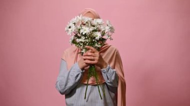 Gorgeous Middle Eastern Muslim woman with head covered in pink hijab, enjoying the smell of a spring bouquet of flowers, smiling looking at camera with an insightful gaze, isolated on pink background