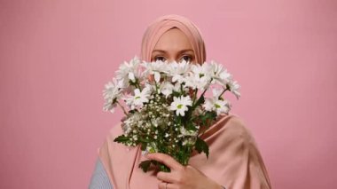 Close-up portrait of beautiful confident Arab Muslim woman wearing pink hijab, looking at camera with insightful gaze, through a cute bouquet of spring flowers, cutely smiling on pink color background