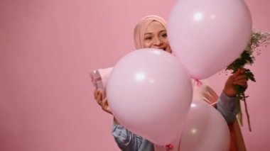 Pembe tesettürlü mutlu Ortadoğulu Müslüman kadın, şişme pembe helyum balonlarının arkasında beliriyor, kalp şeklinde şirin bir hediye kutusu ve bir buket çiçek tutuyor, gülümsüyor, pembe arka planda izole edilmiş.