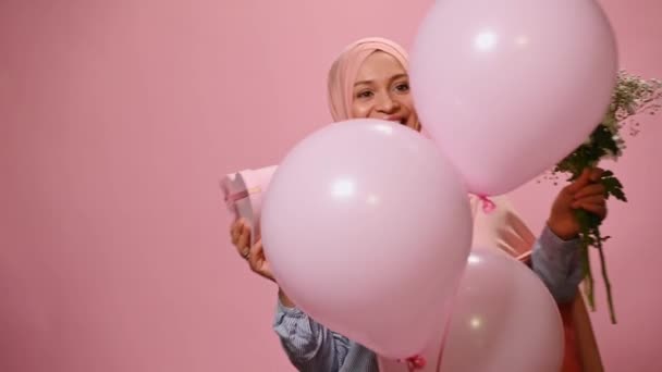 ピンクヒジャーブのハッピー中東イスラム教徒の女性は インフレータブルピンクヘリウム風船の後ろに表示され ハート型のかわいいギフトボックスと花の花束を保持し ピンクの背景に隔離された — ストック動画
