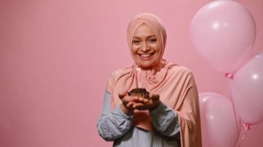 Beautiful modern Arab Muslim woman in stylish pink hijab, making a cherished wish on her birthday and blowing out a candle on the cupcake, posing next to pink inflatable balloons over pink background