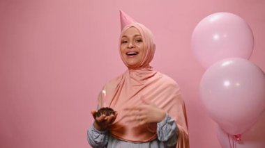 Cheerful Arab Muslim woman wearing a pink hijab and a birthday cap, singing song and holding out at camera a birthday cake with lit candle, smiling, isolated on pink background with helium balloons