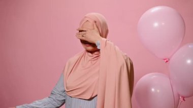 Çekici Müslüman kadın gözlerini kapatıyor, sürpriz bir şekilde doğum günü ya da Kadın Günü hediyesi alıyor, kameraya gülümsüyor, pembe renkli arka planda helyum balonlarıyla.