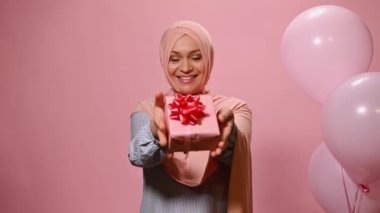 Kafası tesettüre bulanmış, güzel, orta yaşlı, Müslüman bir kadın, kameraya sevimli bir hediye gösteriyor, kameraya dişlek bir gülümseme, helyum balonlarıyla poz veriyor, piknik arka planında izole edilmiş.