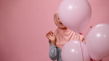 Güzel, mutlu, başı pembe tesettüre bulanmış, neşeli, dişlek gülümseyen, olumlu duygular ifade eden, pembe renkli arka planda pembe şişme balonlarla poz veren Müslüman bir kadın.
