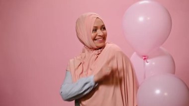 Çekici Orta Doğulu Müslüman kadın, mavi günlük gömlek ve pembe başörtüsü giyiyor, kameraya kurnazca gülümsüyor, pembe renkli arka planda pembe şişme helyum balonları ile poz veriyor..