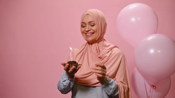 ピンクのヒジャーブの美しい中東イスラム教徒の女性 幸せな誕生日の歌を歌いながらカメラで笑顔 彼女の手の中にライトキャンドルとカップケーキを保持 ピンクの色の背景に隔離 — ストック動画