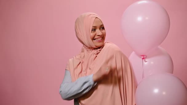 青のカジュアルなシャツとピンクのヒジャーブを身に着けている魅力的な現代的な中東のイスラム教徒の女性は ピンク色の背景にピンクのインフレータブルヘリウム風船でポーズをとって カメラを見てかわいい笑顔 — ストック動画