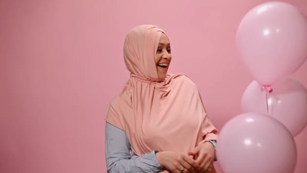 ピンクヒジャーブの魅力的な中年アラブイスラム教徒の女性は ピンクの色の背景にヘリウム風船が現れる間に驚きを表現します 中東の女性は誕生日パーティーを祝い カメラで微笑んだ — ストック動画