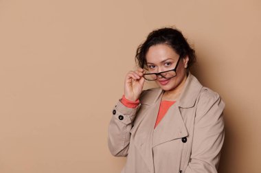 Positive multi-ethnic middle-aged smiling woman in casual wear, puts on stylish eyeglasses, looking at camera over isolated beige background with free space for ads. Ophthalmology. Eye vision concept