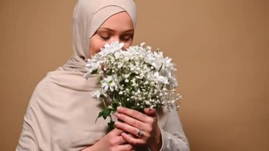 Close-up portrait of gorgeous Middle-Eastern Muslim woman with head covered in beige hijab, enjoying the smell of a spring bouquet of beautiful flowers for Womens Day, on isolated cream background