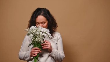 Charming elegant and feminine middle-aged multi-ethnic woman receives flowers, holding beautiful bouquet and smiling at camera, beige background. Valentines day, romance, celebration, beauty concept.
